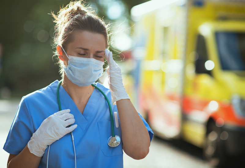 Photo of a healthcare worker wearing scrubs, gloves, and a mask while touching her temple as if she has a headache. Source: U.S. DOL Blog